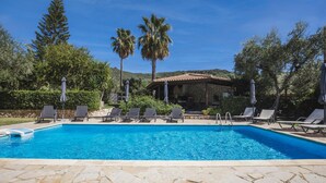 Una piscina cristallina con lettini e ombrelloni, circondata da alberi e un edificio in pietra.