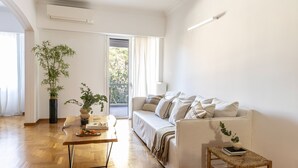 A modern living room with a wooden floor, a white sofa, a small table with a plant, and a view of the outdoors through a door. - Acropolis Museum Retreat - 2 Bedroom Apartment (Athina)