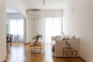 A spacious living room with a wooden floor, a white sofa, a small table with a plant, and a dining area with a chair and table. - Acropolis Museum Retreat - 2 Bedroom Apartment (Athina)