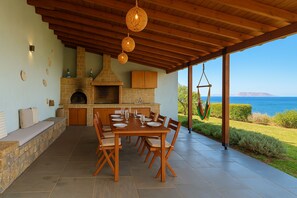 An outdoor kitchen with a stone fireplace, wooden dining table, and chairs, overlooking a scenic view of the sea. - Luxury  Villa  Kopsas (Kokkini Hani)