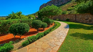 A stone pathway leading through a landscaped garden with neatly trimmed bushes and trees, a stone wall, and a mountain in the background. - Luxury  Villa  Kopsas (Kokkini Hani)