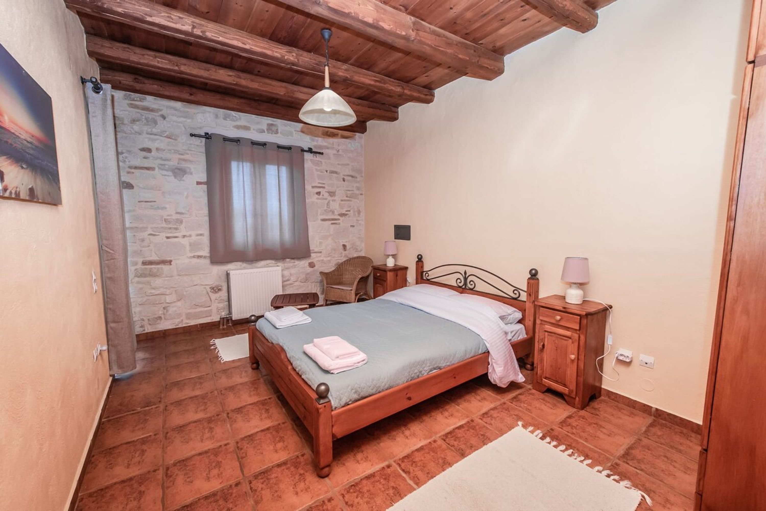 Une chambre avec un mur en pierre, des poutres apparentes en bois au plafond, un lit recouvert d’une courtepointe bleue, une table de chevet avec une lampe et une fenêtre avec des rideaux.
