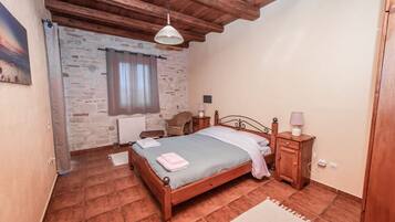 Une chambre avec un mur en pierre, des poutres apparentes en bois au plafond, un lit recouvert d’une courtepointe bleue, une table de chevet avec une lampe et une fenêtre avec des rideaux.