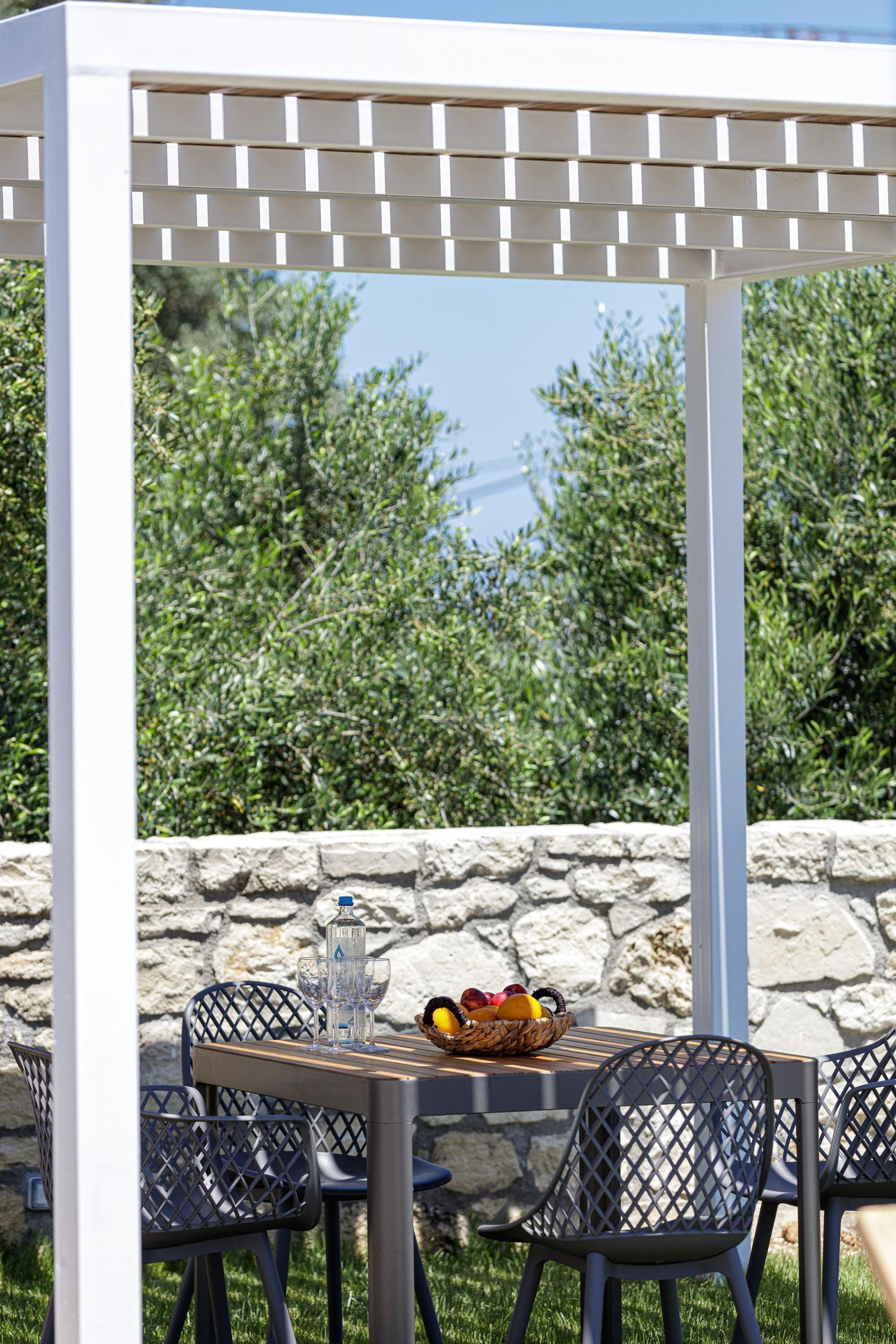 An outdoor dining area with a pergola, stone wall, and greenery in the background.