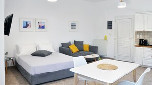 Een moderne, goed verlichte kamer met een bed, een bank en een eettafel. Er hangen ingelijste foto’s aan de muur, er is een televisie en een keukenhoek met kastjes en een radiator.