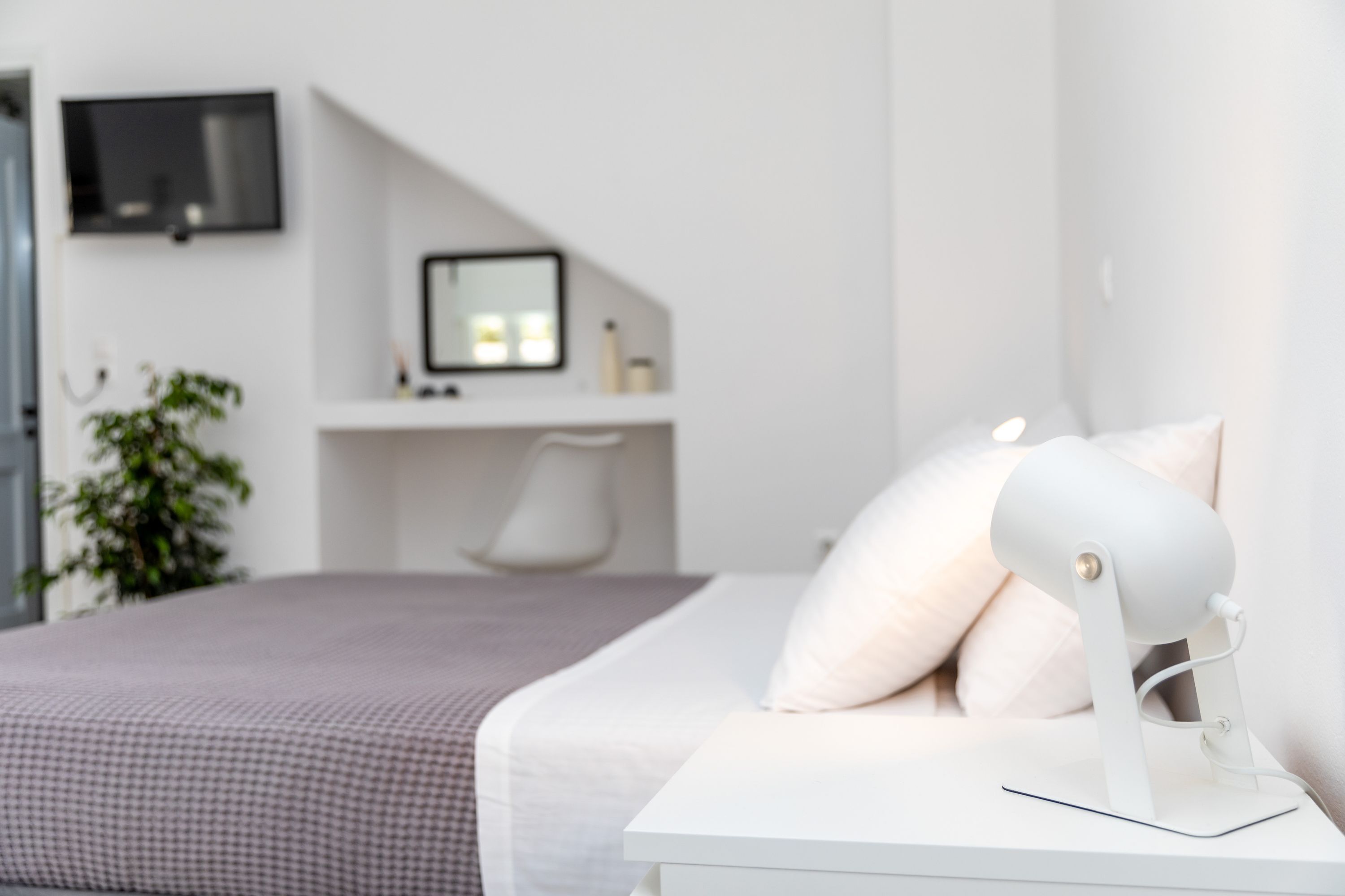 A modern bedroom with a bed, a bedside table with a lamp, a desk with a computer monitor, and a TV mounted on the wall.