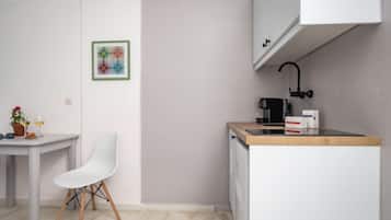 A small kitchenette with a white countertop, a coffee machine, and a sink. There is a white chair and a table with a potted plant and a glass of wine.