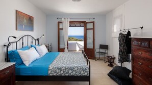 Een slaapkamer met een bed, een dressoir, een stoel en uitzicht op de zee door een open raam.