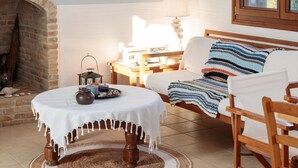 Een gezellige woonkamer met een open haard, een ronde tafel met een witte tafelkleed, een bank met een gestreepte plaid en een groot raam met houten kozijnen.