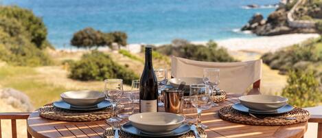 A round wooden table set for dining with plates, glasses, and a bottle, overlooking a beach with the ocean and rocky landscape.