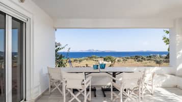 A covered patio with a table and chairs, overlooking a coastal landscape with mountains in the distance.
