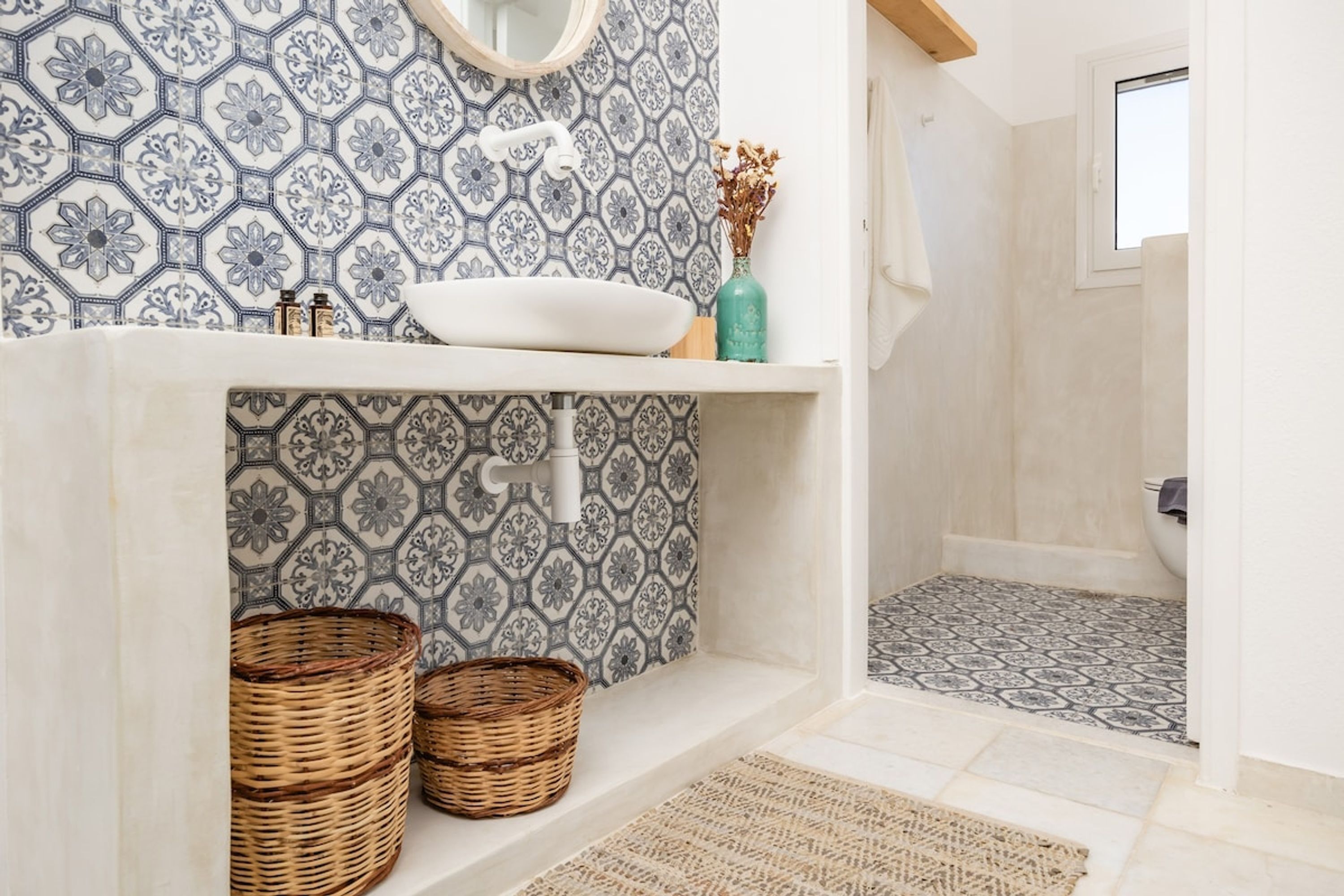 A bathroom with a white sink, a mirror, and a patterned tile backsplash. There are wicker baskets and a woven mat on the floor.