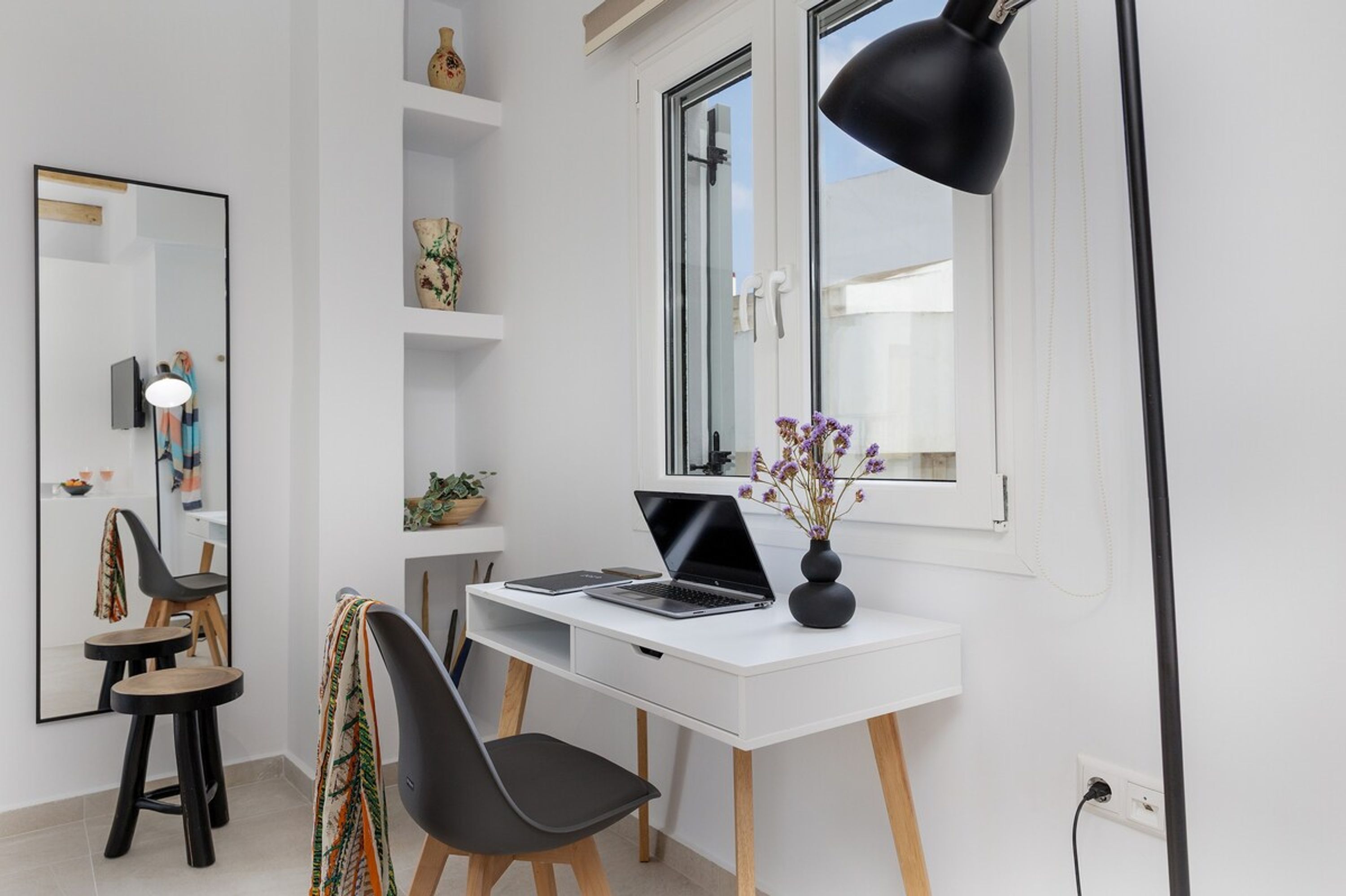 A modern office space with a white desk, black chair, and a laptop. There is a large mirror on the wall, a window with a black desk lamp, and shelves with decorative items.