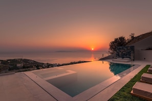 A modern outdoor pool area with a clear view of the sunset over the sea, a stone wall, and lounge chairs. - Luxury   Villa   Vie (Agios Dimitrios Rodias)