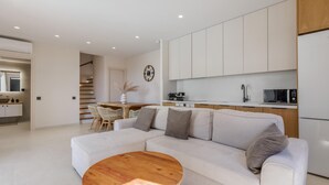 Una sala de estar moderna con un sofá blanco, una mesa de centro de madera y una zona de cocina con armarios y electrodomésticos blancos.