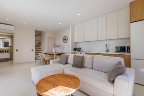 A modern living room with a white sofa, a wooden coffee table, and a kitchen area with white cabinets and appliances. - Cameo 2  Luxury Villa with Pool (Zakynthos)