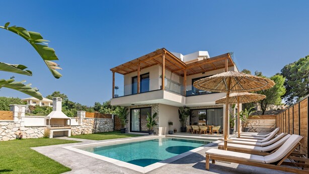 Una casa moderna de dos pisos con piscina, mobiliario exterior y un muro de piedra.