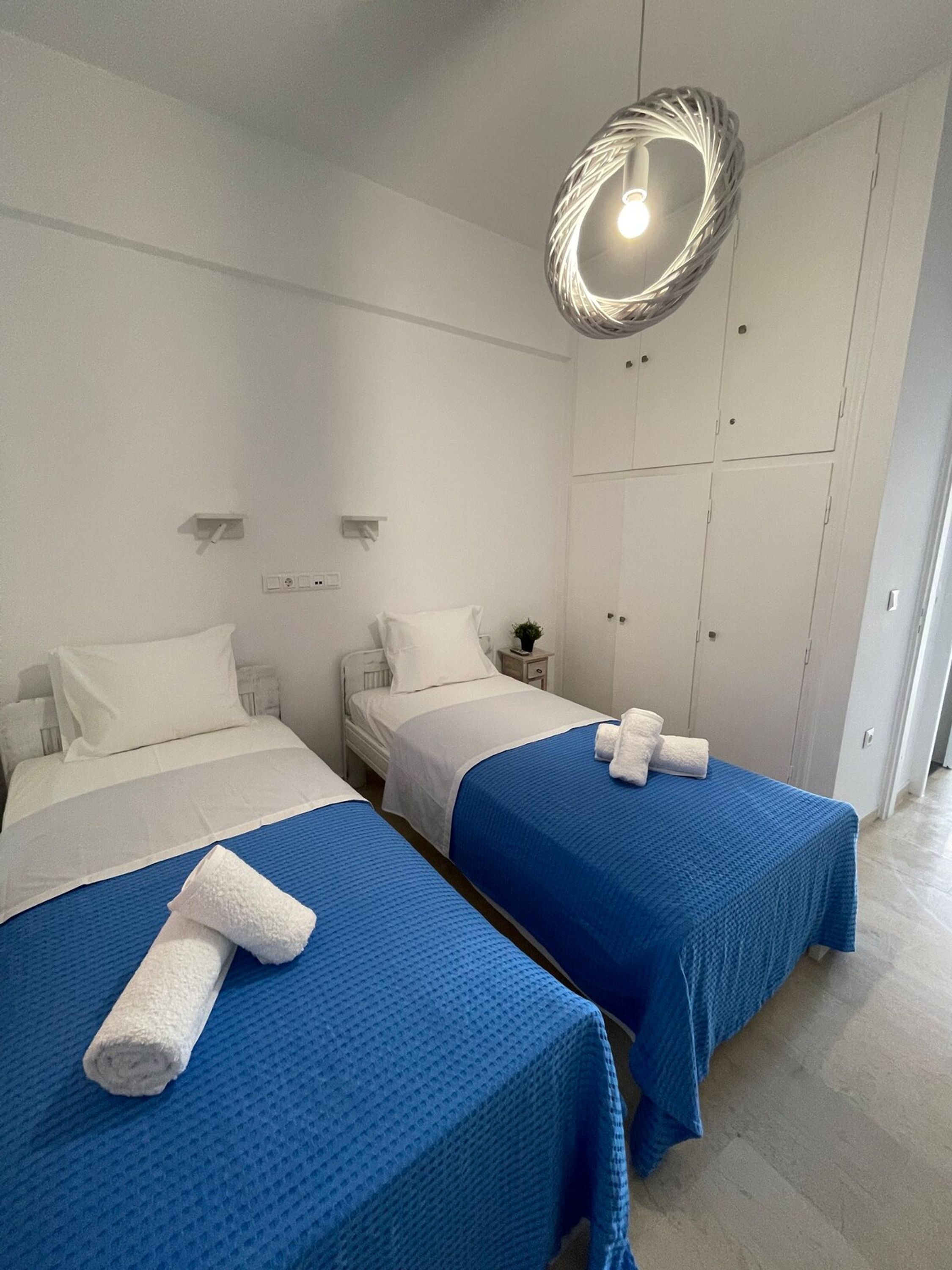 A hotel room with two beds, each with a blue bedspread and white pillows. There is a white wardrobe with a circular mirror above the door, and a small bedside table with a lamp and a plant.