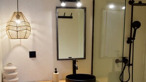 Ein modernes Badezimmer mit einem Holzbadezimmerhocker, einem runden schwarzen Waschbecken, einem großen Spiegel, einer hängenden Leuchte und einer Duschkabine.