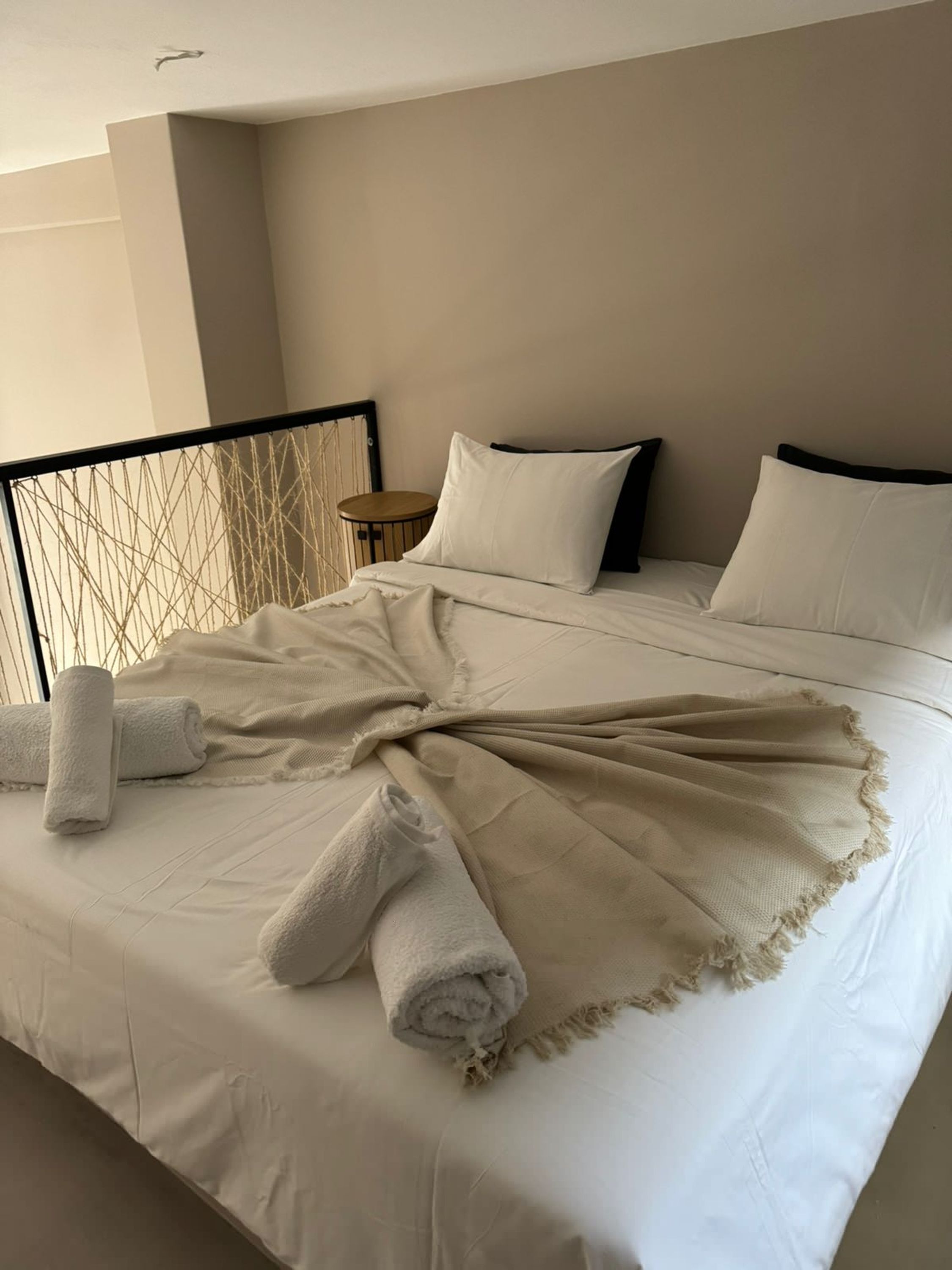 A neatly made bed with white linens and rolled towels, a round wooden side table, and a headboard with a geometric pattern.