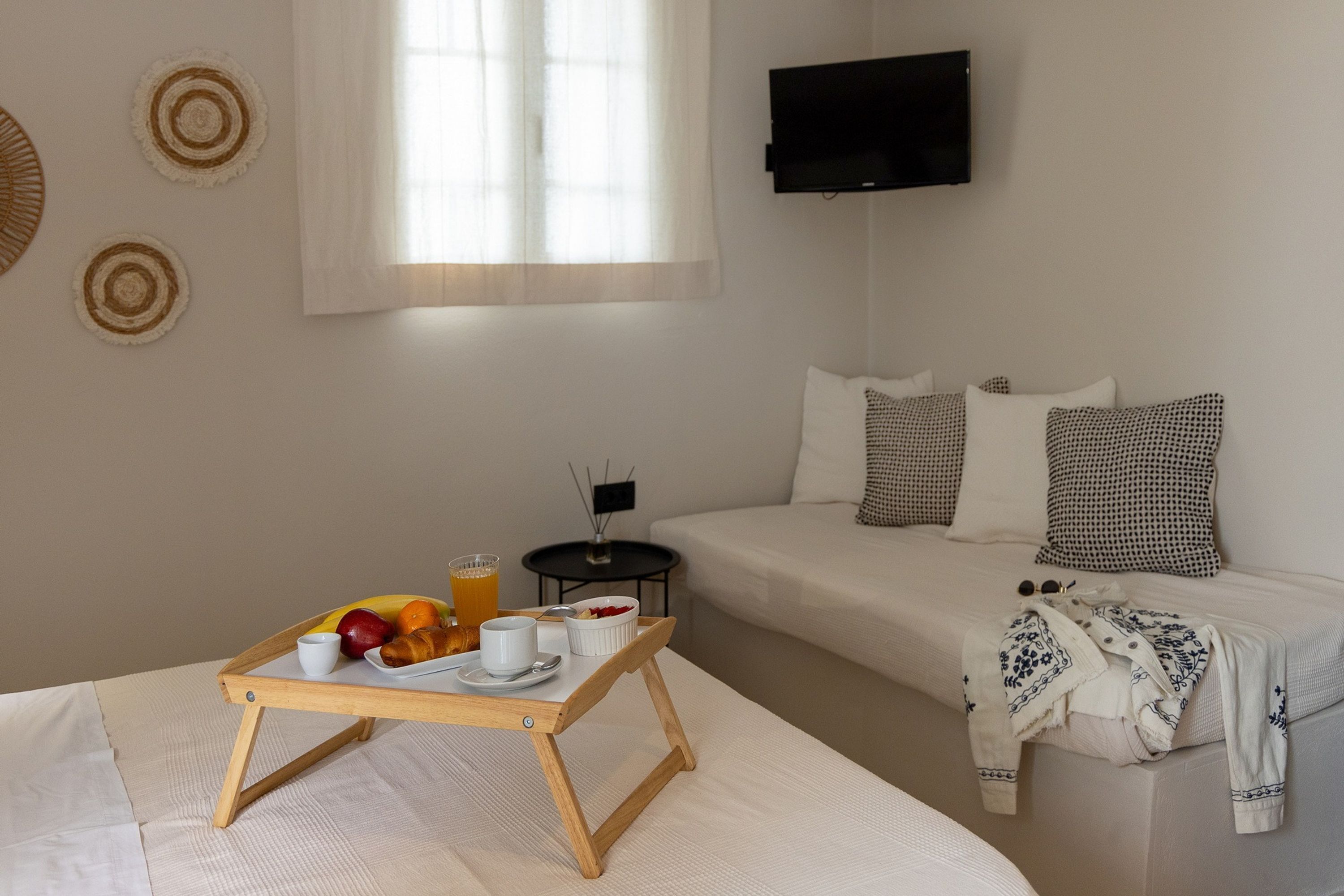 Ein ordentlich gedeckter Frühstückstisch mit Tablett, Obstschale, Tasse und Glas Orangensaft. Im Hintergrund ist ein Bett mit weißer Decke und gemusterten Kissen zu sehen.