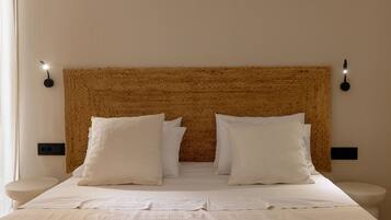 Uma cama bem arrumada com lençóis e almofadas brancas, um painel de cabeceira tecido com textura e mesas de cabeceira com luminárias pretas fixadas na parede.