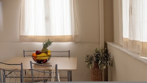 A dining area with a table, chairs, a fruit bowl, a potted plant, and sheer curtains. - Salt & Stone In Center Of Tinos Luxury Suite (Tinos)