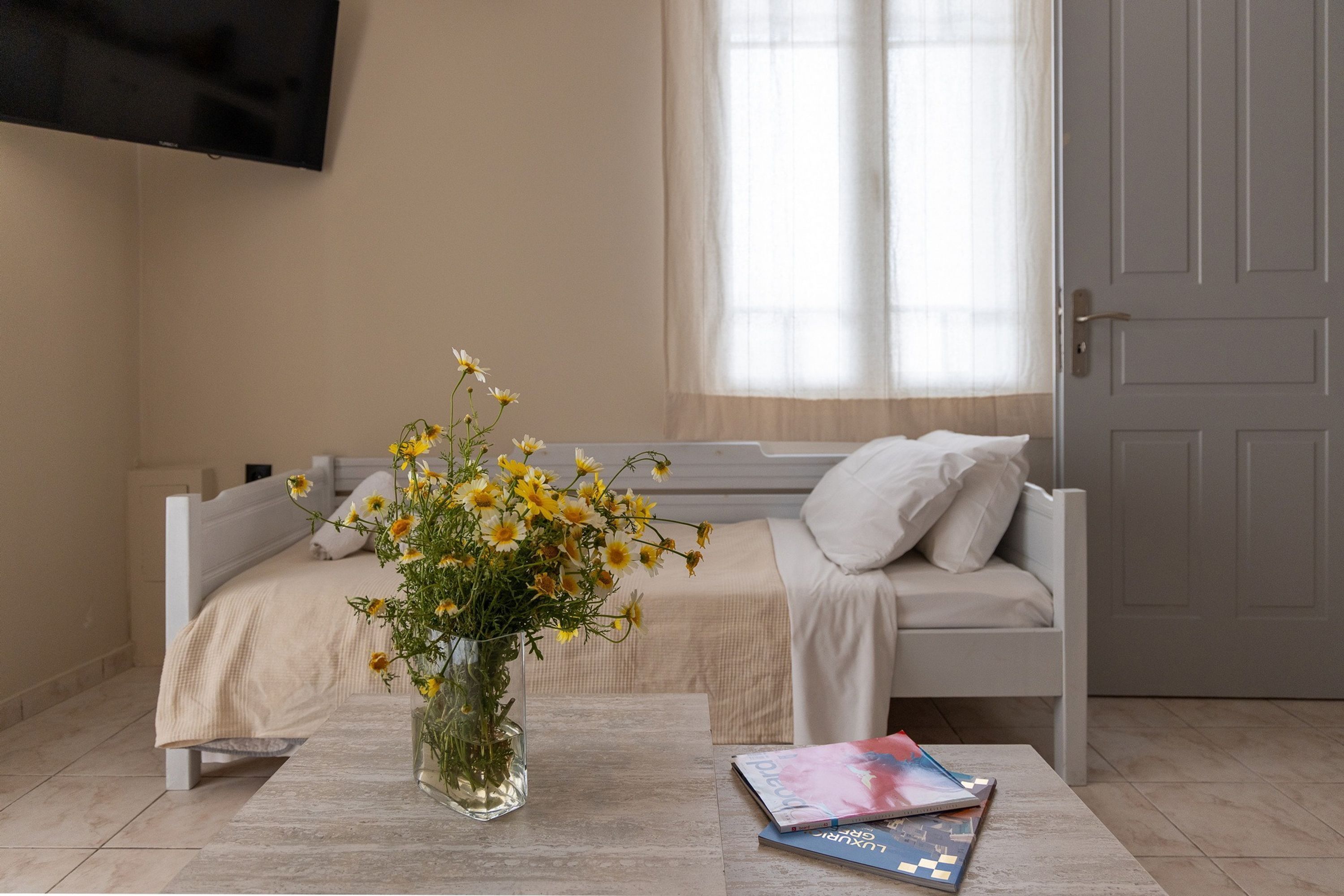 Ein Schlafzimmer mit einem Bett, einem Nachttisch mit Blumenvase und einem Fenster mit transparenten Vorhängen.