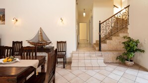 A dining area with a wooden table and chairs, a decorative chair, a small table with fruit, and a potted plant near a staircase. - Villa Lefki Limni Retreat 300m from beach (Corfu)