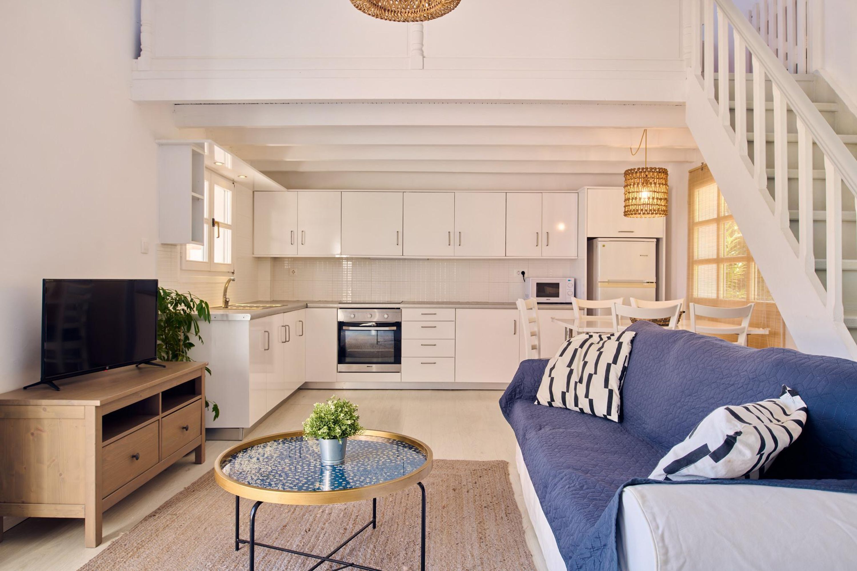 A modern living room with a sofa, a coffee table, and a TV. There is a kitchen in the background with white cabinets and a dining area with chairs.