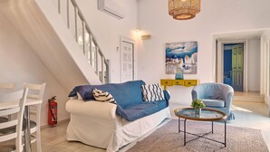 Un salon moderne avec un escalier blanc, un canapé bleu, une table basse ronde et un tableau représentant un paysage urbain.