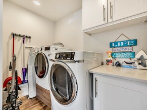 A laundry room with two front-loading washing machines, a drying rack, a vacuum cleaner, and a shelf with cleaning supplies.