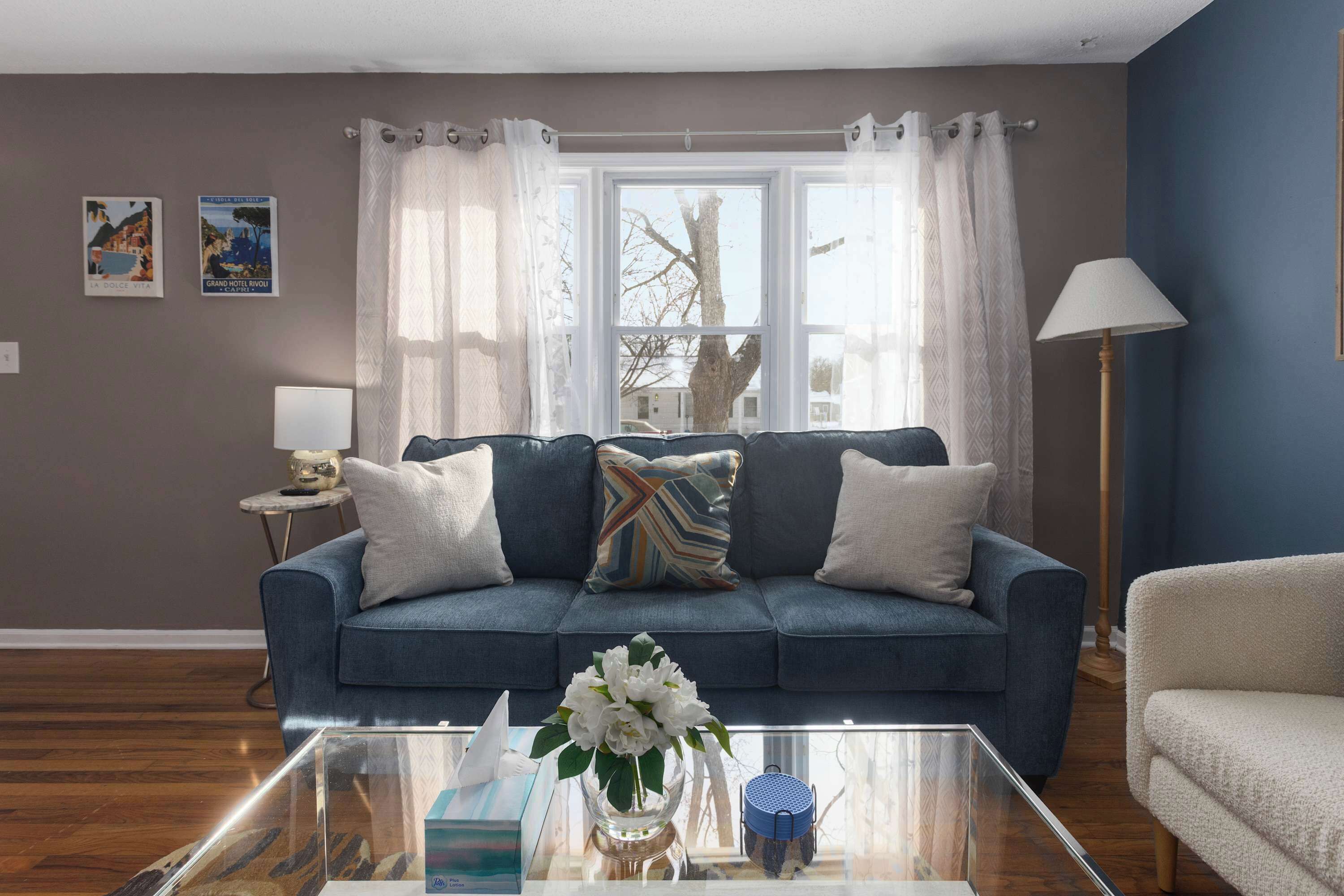 Una sala de estar con un sofá azul, una mesa de centro de vidrio y un jarrón con flores. Hay dos almohadones en el sofá, uno con estampado a rayas. Una lámpara está sobre una mesita auxiliar al lado del sofá.