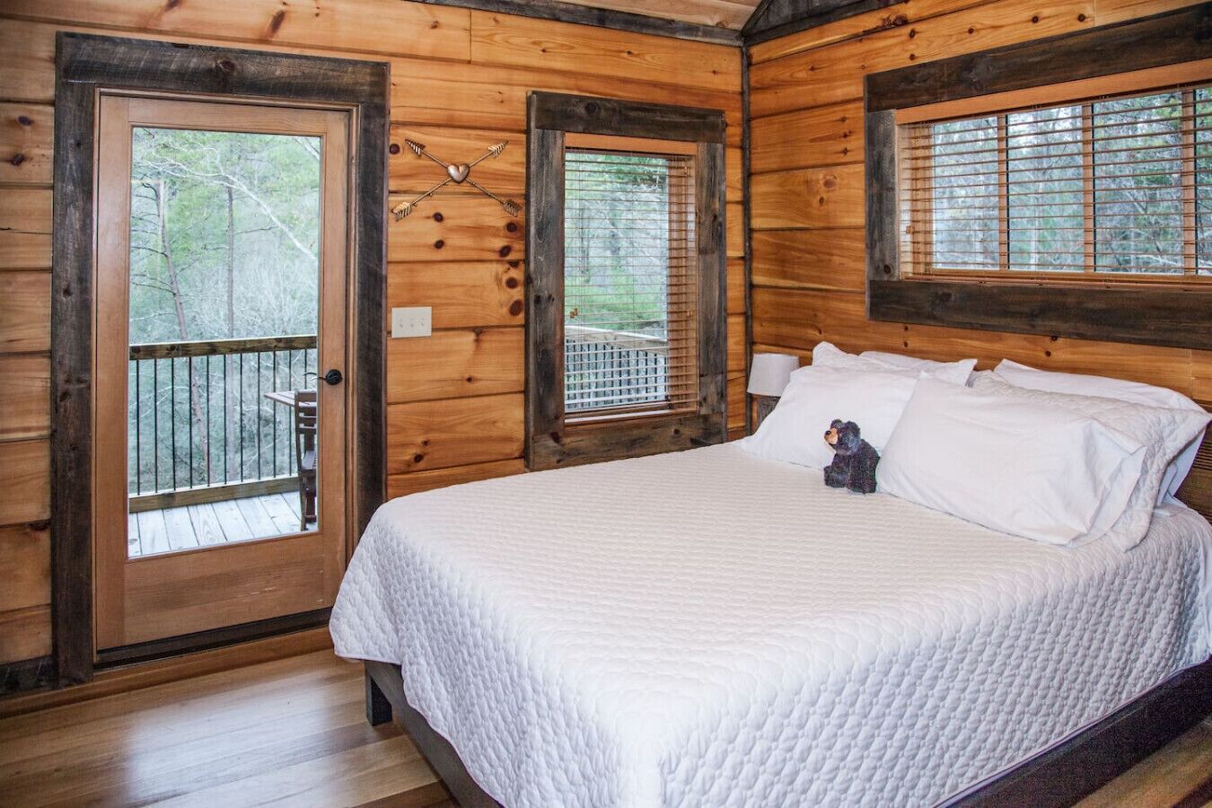 A wooden cabin bedroom with a bed, a small bear decoration, and a balcony with a view of trees.