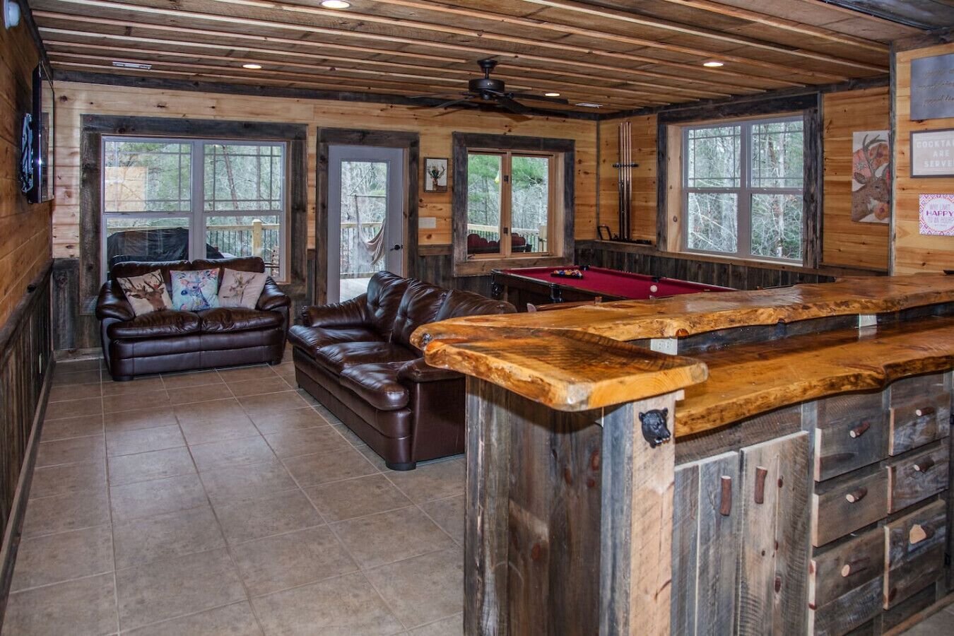 A rustic cabin interior with a wooden ceiling, a pool table, a leather sofa, and a bar area.