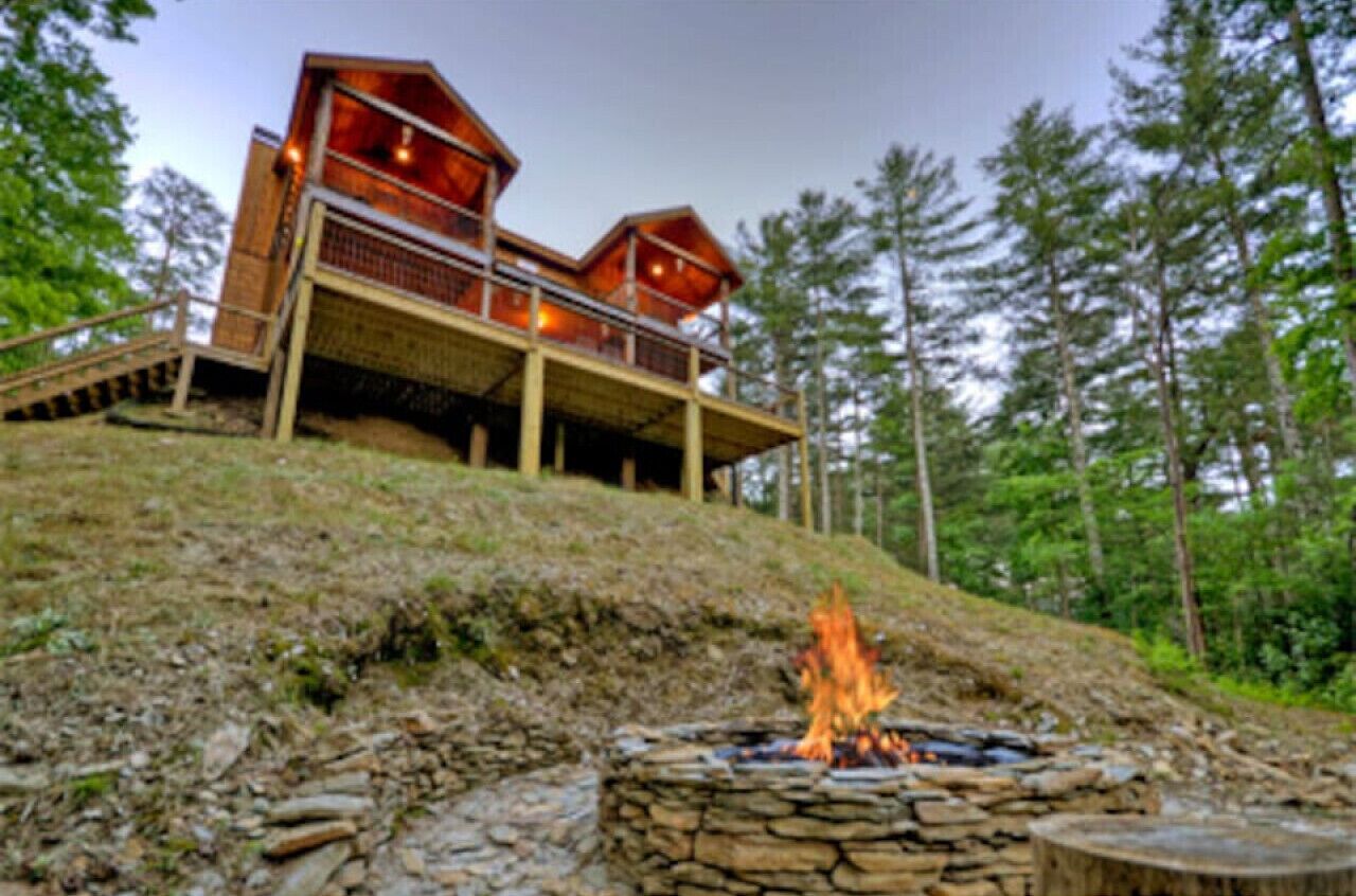 A modern wooden cabin with a stone fire pit, surrounded by a forest.