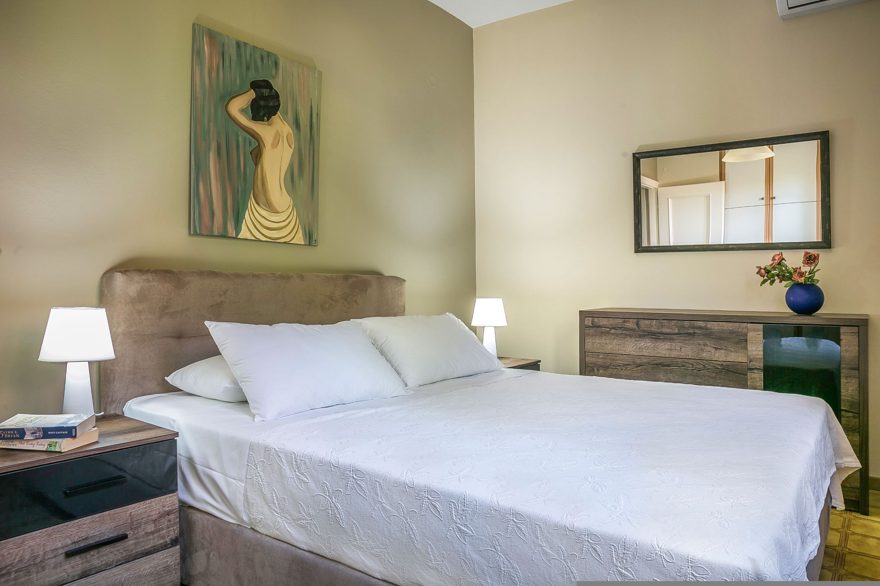 A neatly made bed with white linens, a headboard, bedside tables with lamps, a wooden nightstand, a dresser, a mirror, and a painting on the wall.
