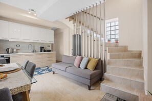 A modern kitchen with white cabinets, a dining table with chairs, and a gray sofa with colorful cushions. A staircase with a wooden railing leads to a landing with a window. - Park and sea view Villa with Private Pool 2 (Chania)