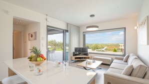 A modern living room with a sofa, a coffee table, a TV, and a large window offering a view of the outdoors. - Sunset Villa in Moudania (Moudania)