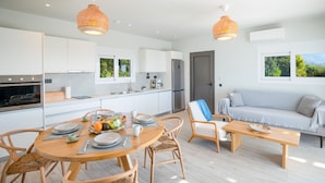 Cozinha moderna com armários brancos, sala de jantar com mesa de madeira redonda e um sofá com vista para vegetação.