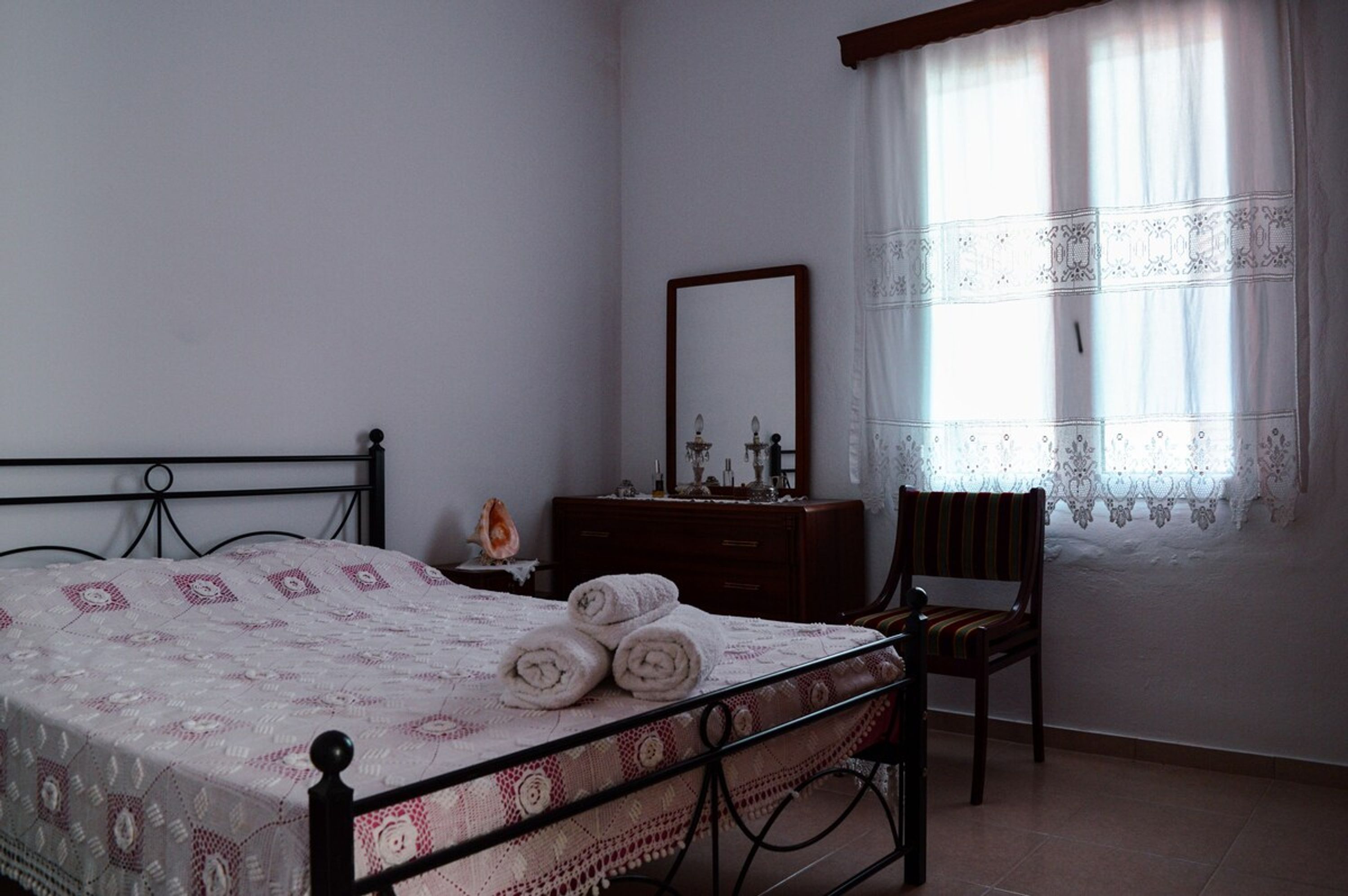 Un dormitori amb llit, una petita estanteria de fusta, una cadira, un mirall i una finestra amb cortines.