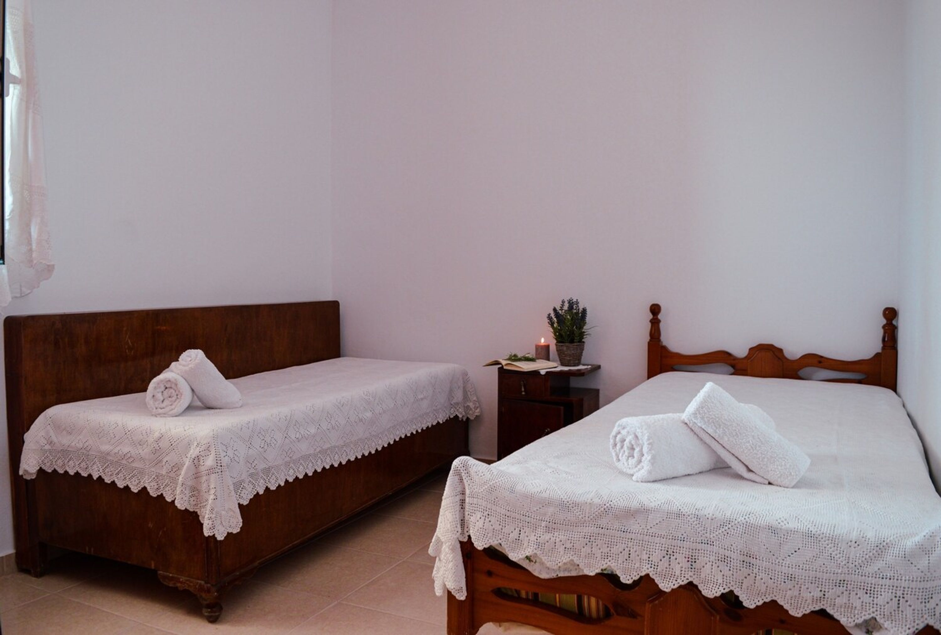 Dos camas de madera con sábanas blancas y cubrecolchas de encaje, una mesita de noche con una planta y una ventana con cortina.