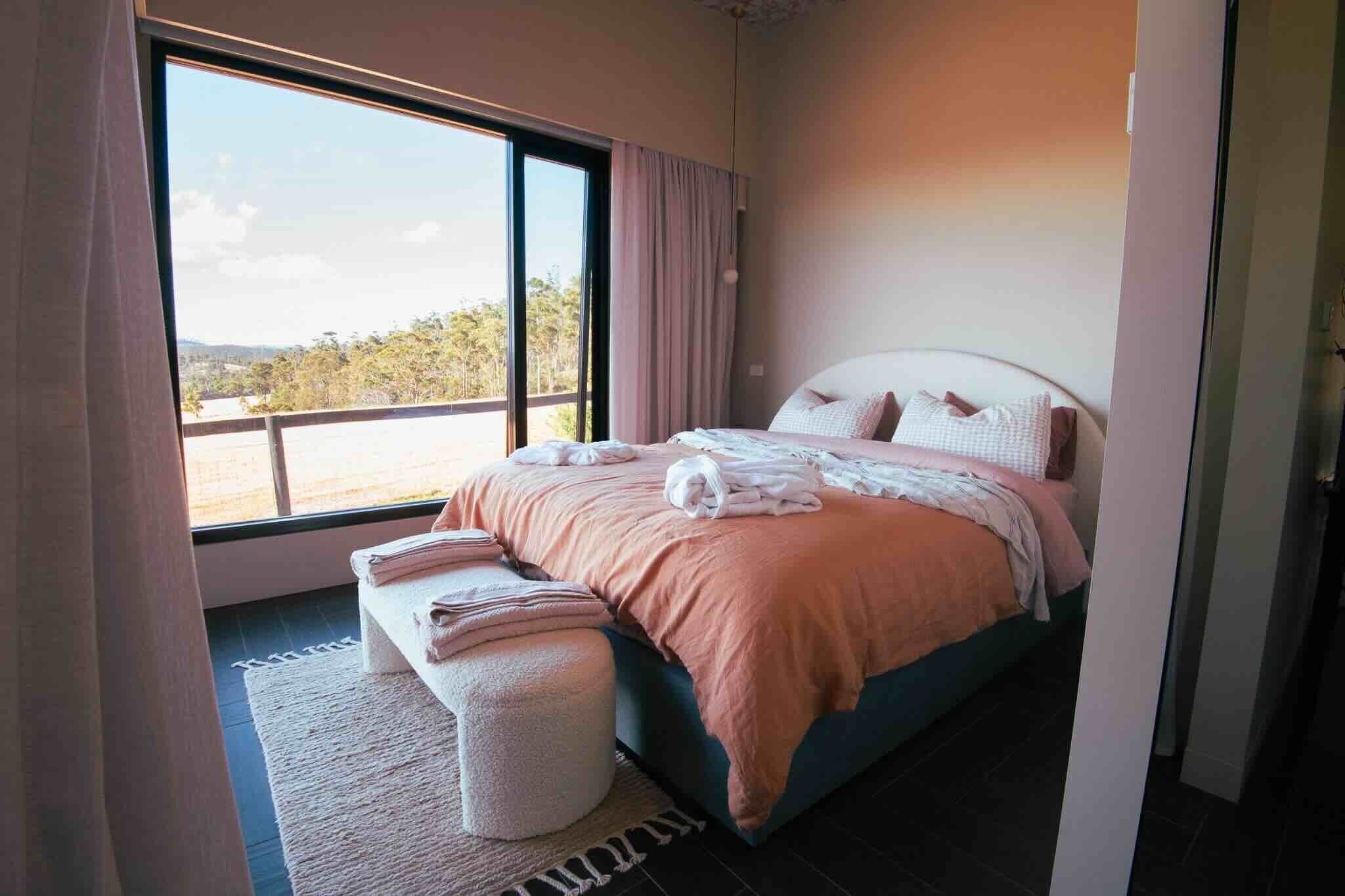 Ein Schlafzimmer mit großem Fenster, einem Bett mit Kissen und Decke sowie einem kleinen Hocker.