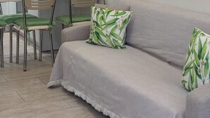A modern living room with a grey sofa, green cushions, a dining area with chairs, a kitchen with white cabinets, and a wall-mounted light fixture. - Pistachio Suite - Town Centre (Kerkira)