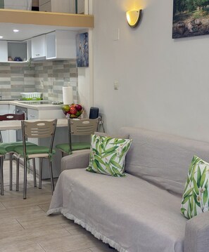 A modern living room with a grey sofa, green cushions, a dining area with chairs, a kitchen with white cabinets, and a wall-mounted light fixture. - Pistachio Suite - Town Centre (Kerkira)