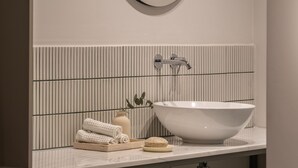 A modern bathroom with a large round mirror, a white sink, and striped tile backsplash. - Iris - By Celestial Suites (Athina)