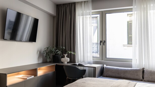 A modern hotel room with a bed, a desk with a chair, a TV mounted on the wall, and a window with curtains. - Nyx - by Celestial Suites (Athina)