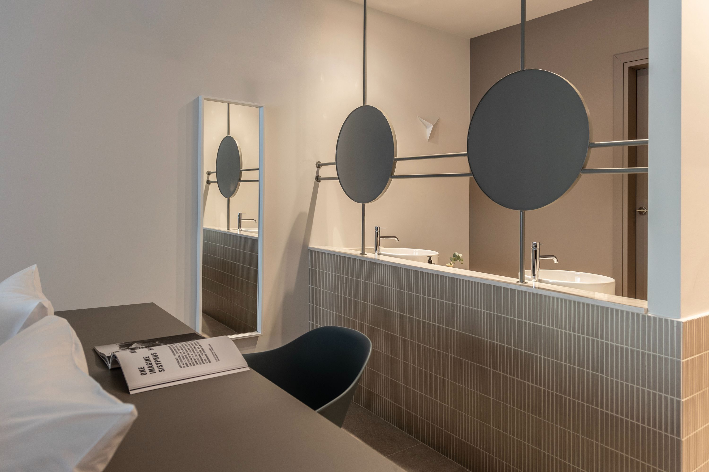 A modern bathroom with a large mirror, a round mirror, and a sink. There is a chair and a table with a magazine.