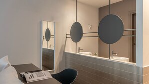 Ein modernes Badezimmer mit einem großen Spiegel, einem runden Spiegel und einem Waschbecken. Ein Stuhl und ein Tisch mit einer Zeitschrift stehen bereit.