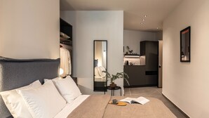 Ein modernes Hotelzimmer mit einem ordentlich bezogenen Bett, einem Nachttisch mit einem Buch und einer Tasse, einem Spiegel sowie Blick ins Bad und die Küchenzeile.